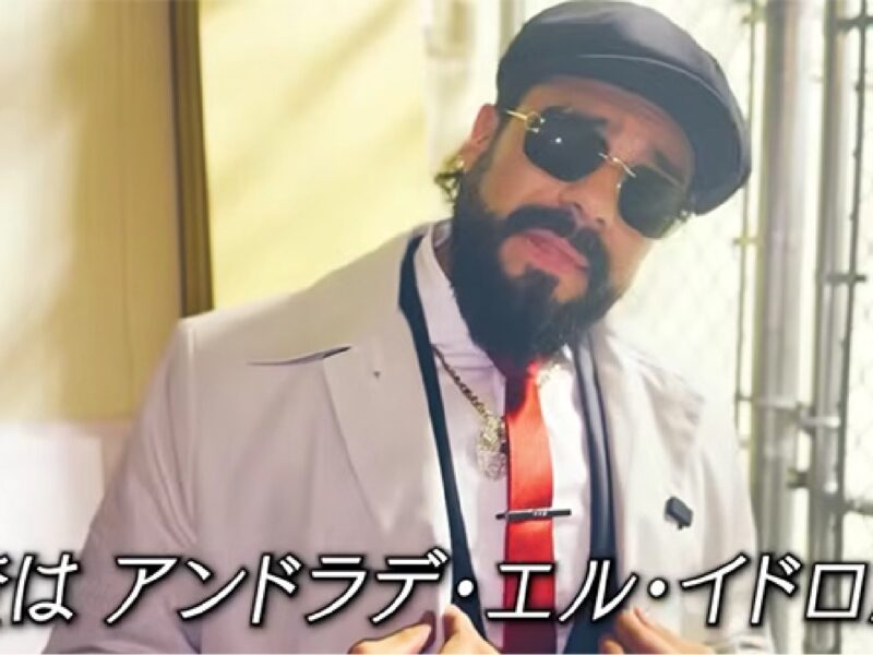 Bearded man in a white suit with a red tie, wearing sunglasses and a cap, posing near a chain-link fence with Japanese subtitles at the bottom.
