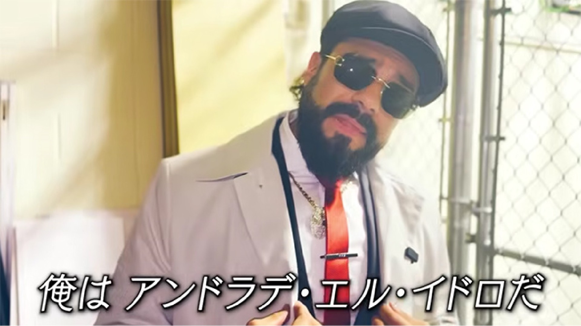 Bearded man in a white suit with a red tie, wearing sunglasses and a cap, posing near a chain-link fence with Japanese subtitles at the bottom.