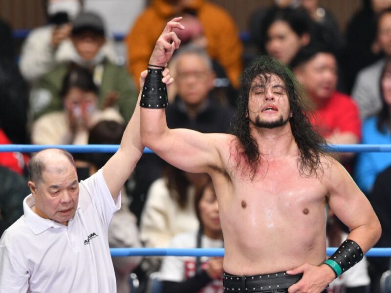 Wrestler with long dark hair and studded wristbands raises his arm in victory as referee holds it up inside the ring.