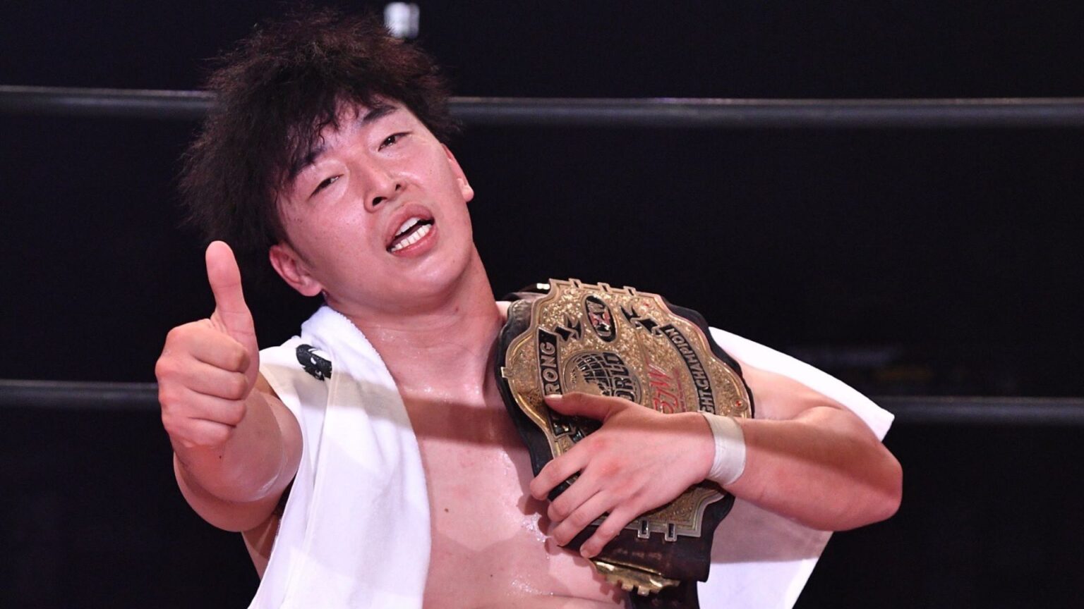 Wrestler with a gold championship belt over shoulder, giving a thumbs-up to the camera