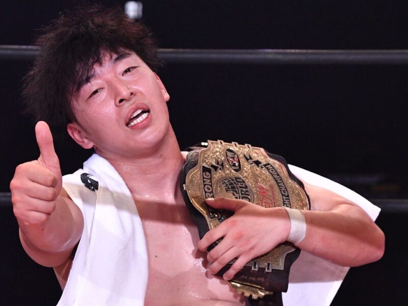 Wrestler with a gold championship belt over shoulder, giving a thumbs-up to the camera