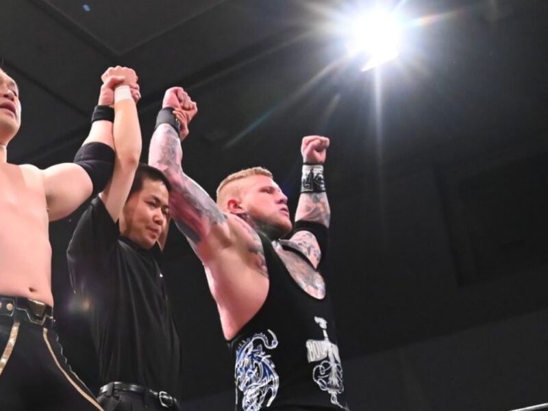 Wrestlers celebrate victory with arms raised in the ring, backed by a referee.