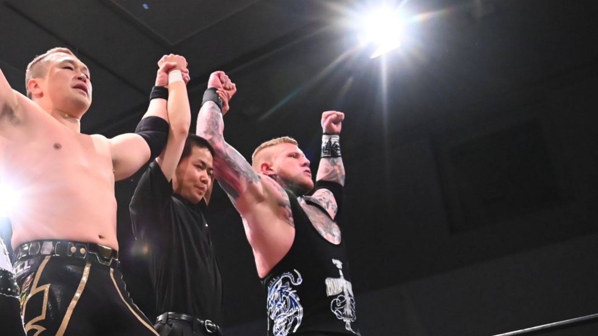 Wrestlers celebrate victory with arms raised in the ring, backed by a referee.