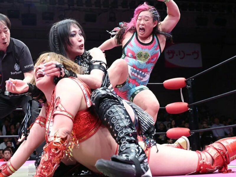 Wrestling match in a ring with four competitors; a wrestler in red gear is on the mat while others grapple around her and a referee watches closely.