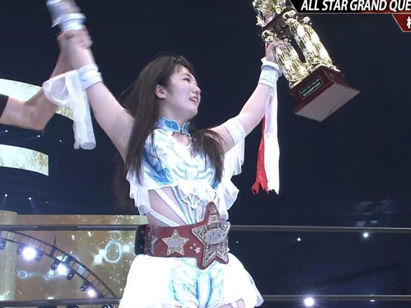 Female wrestler raises large trophy in victory pose on stage, wearing championship belt and white-blue outfit with armbands.