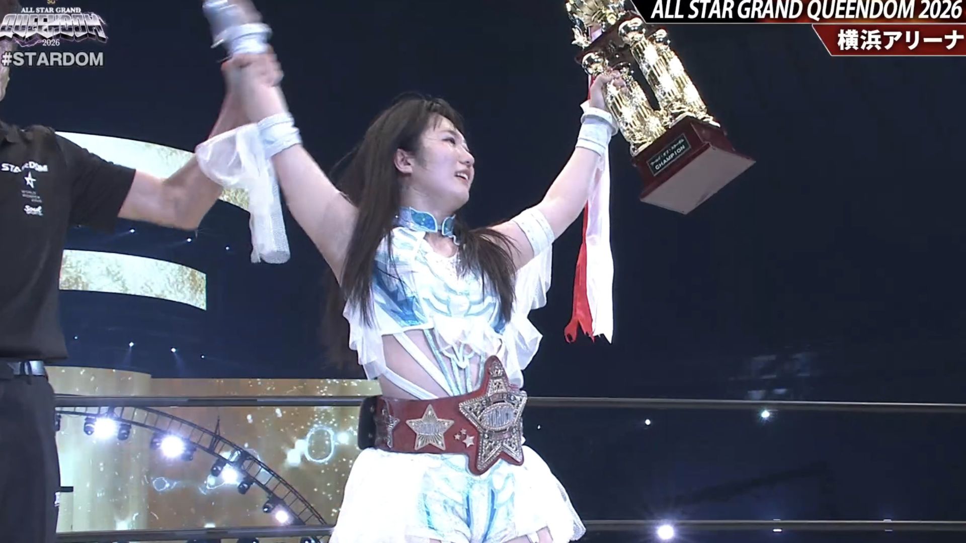Female wrestler raises large trophy in victory pose on stage, wearing championship belt and white-blue outfit with armbands.