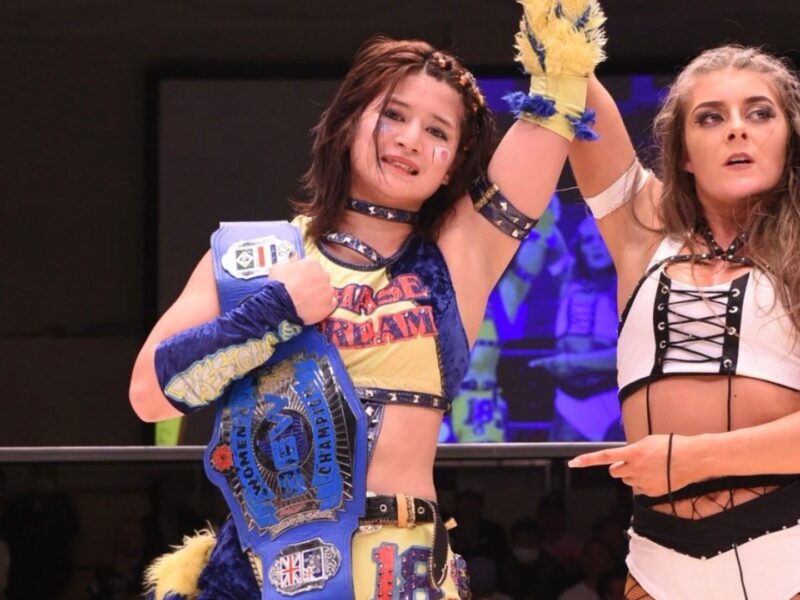 Two female wrestlers celebrate in a ring, one wearing a blue championship belt and colorful outfit with raised arm, the other in a white outfit with black lacing watching nearby.