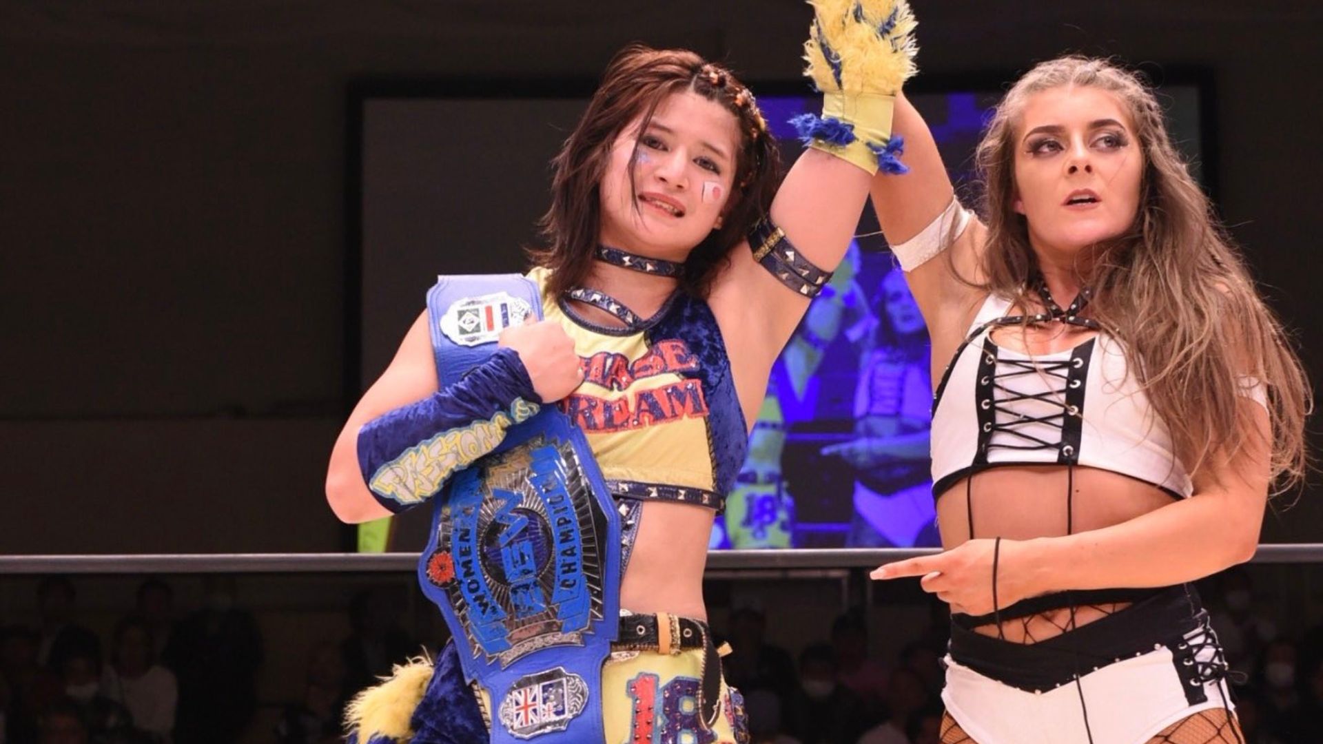 Two female wrestlers celebrate in a ring, one wearing a blue championship belt and colorful outfit with raised arm, the other in a white outfit with black lacing watching nearby.