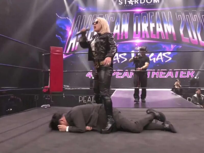 Wrestler in black leather jacket stands with a microphone while another wrestler lies face down on the ring floor in front of a lit backdrop that says 'American Dream 2023.'