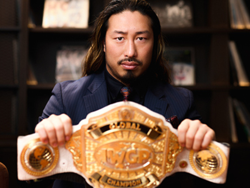 Man in a navy pinstripe suit holds a large gold WGP Championship belt toward the camera with a bookcase background.