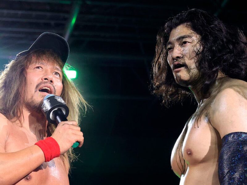 Two muscular wrestlers in a ring during a promo, one holding a microphone and wearing a black cap, the other with long hair and a blue arm sleeve facing him.