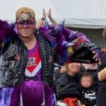 Drag performer in bright, feathered purple costume and bold makeup raises arms at a parade.