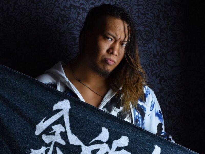 Man with long hair and goatee holds a dark banner with white kanji, against a patterned wall.