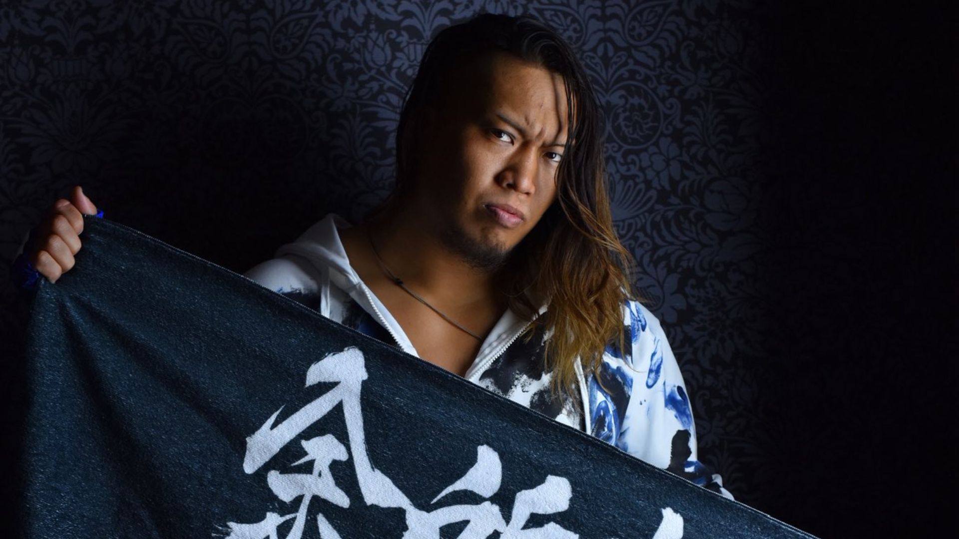 Man with long hair and goatee holds a dark banner with white kanji, against a patterned wall.