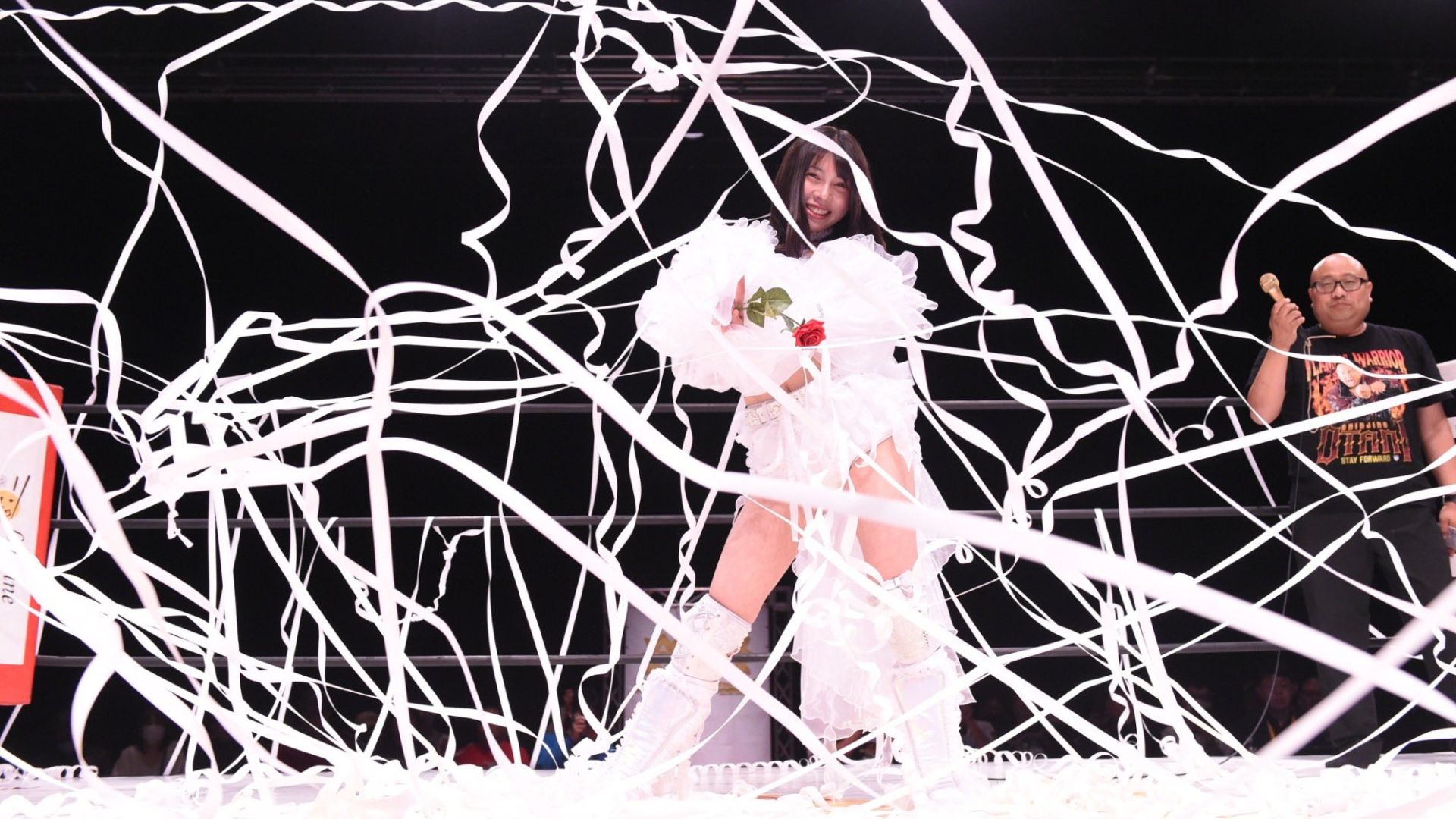 Performer in a white frilly costume standing in a ring, holding a rose, surrounded by white streamers with a smiling crowd in the background.”