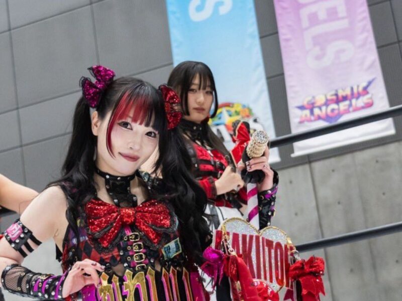 Smiling cosplayer in a red-and-black Gothic outfit with a large bow, posing at a convention with another cosplayer holding a glittery staff in the background.