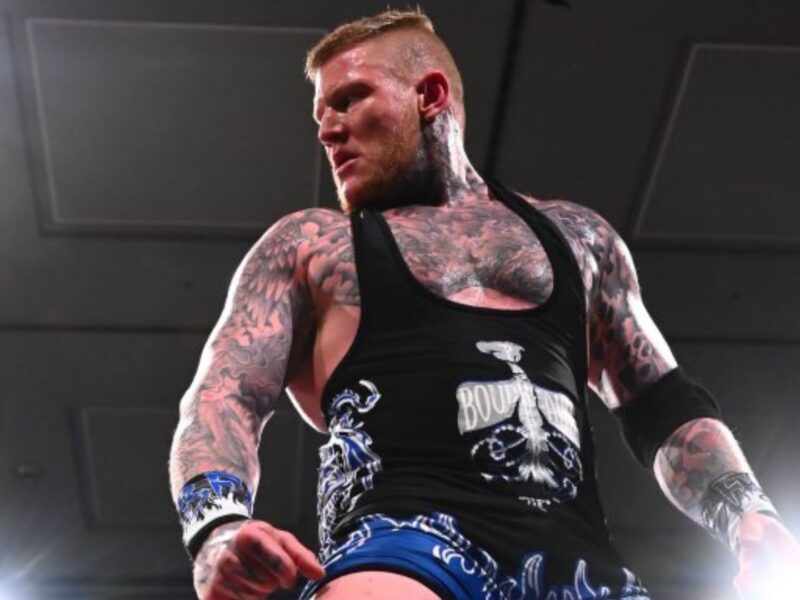 Tattooed male wrestler wearing a black tank top stands in a wrestling ring, ready for action.