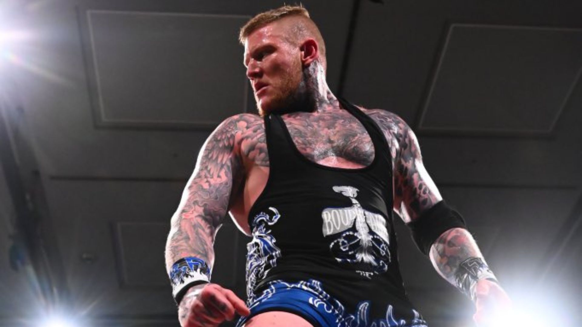 Tattooed male wrestler wearing a black tank top stands in a wrestling ring, ready for action.