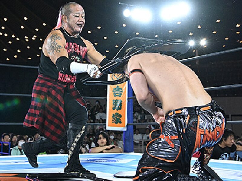 Two pro wrestlers clash in a ring: one in a black sleeveless top and red plaid pants delivering a strike to the other wrestler who is kneeling, with an audience watching in the background.