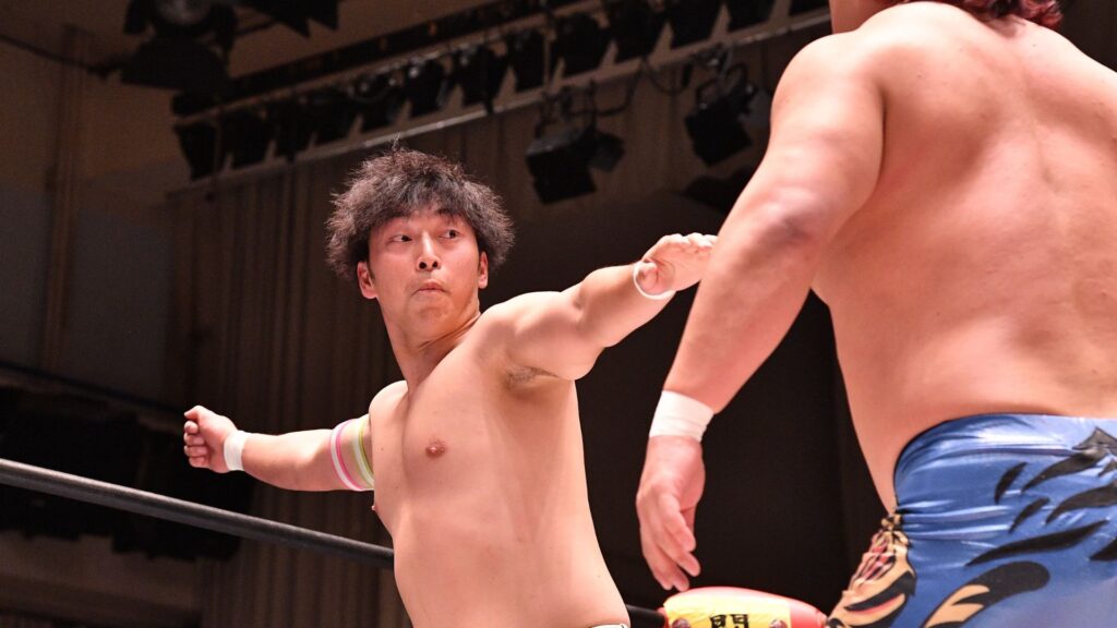 Two shirtless male wrestlers in a ring, one with an outstretched arm toward the other as they clash in a match.