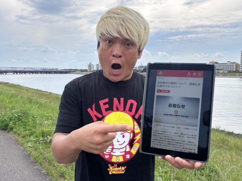 Man with bleached blond hair in a black T‑shirt holds a tablet showing a Japanese notice beside a river and bridge in the background.