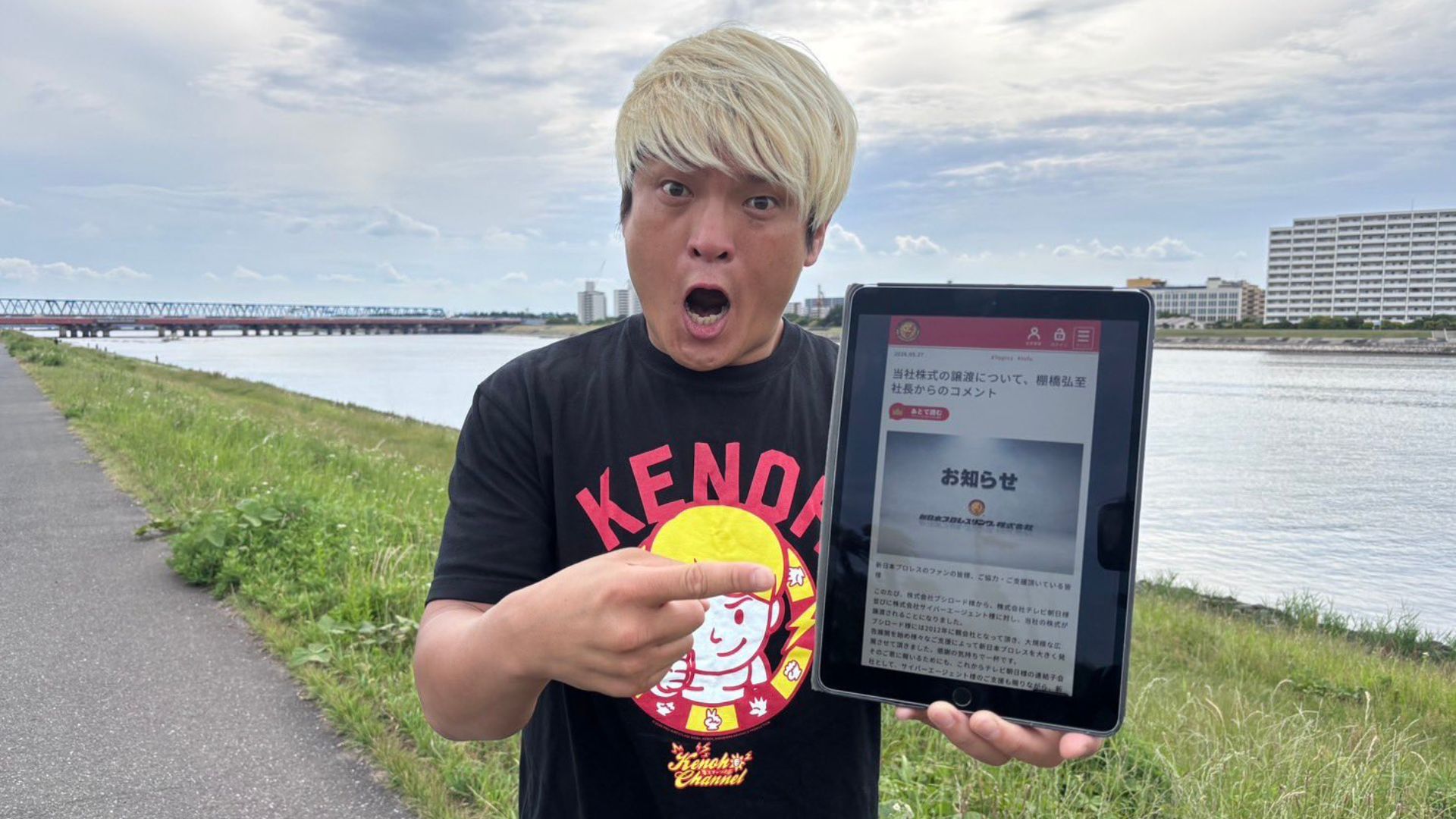 Man with bleached blond hair in a black T‑shirt holds a tablet showing a Japanese notice beside a river and bridge in the background.