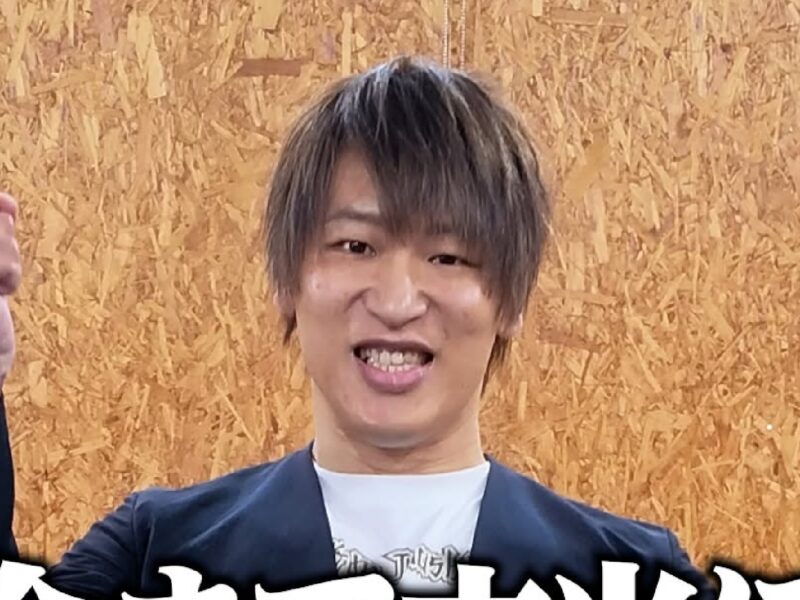Young man with gray-streaked hair smiles and raises a clenched fist in celebration against a plywood background.