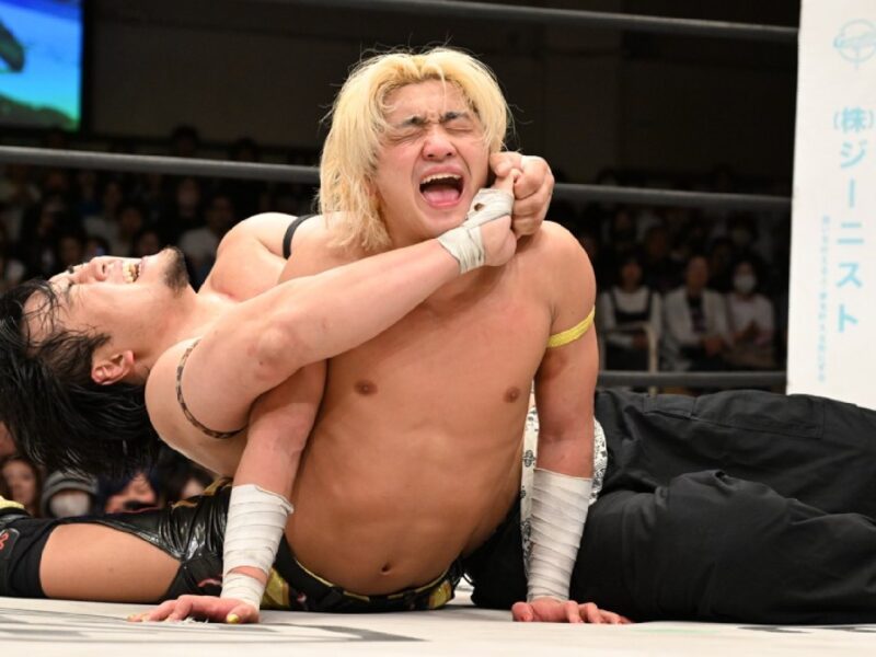 Wrestler with blond hair applies chokehold on a wearing-black tights opponent in a wrestling ring while crowd watches in the background.
