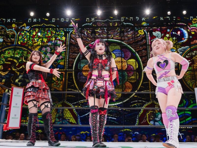 Three female wrestlers pose in a colorful ring with a stained-glass backdrop, bright costumes and dramatic gestures.