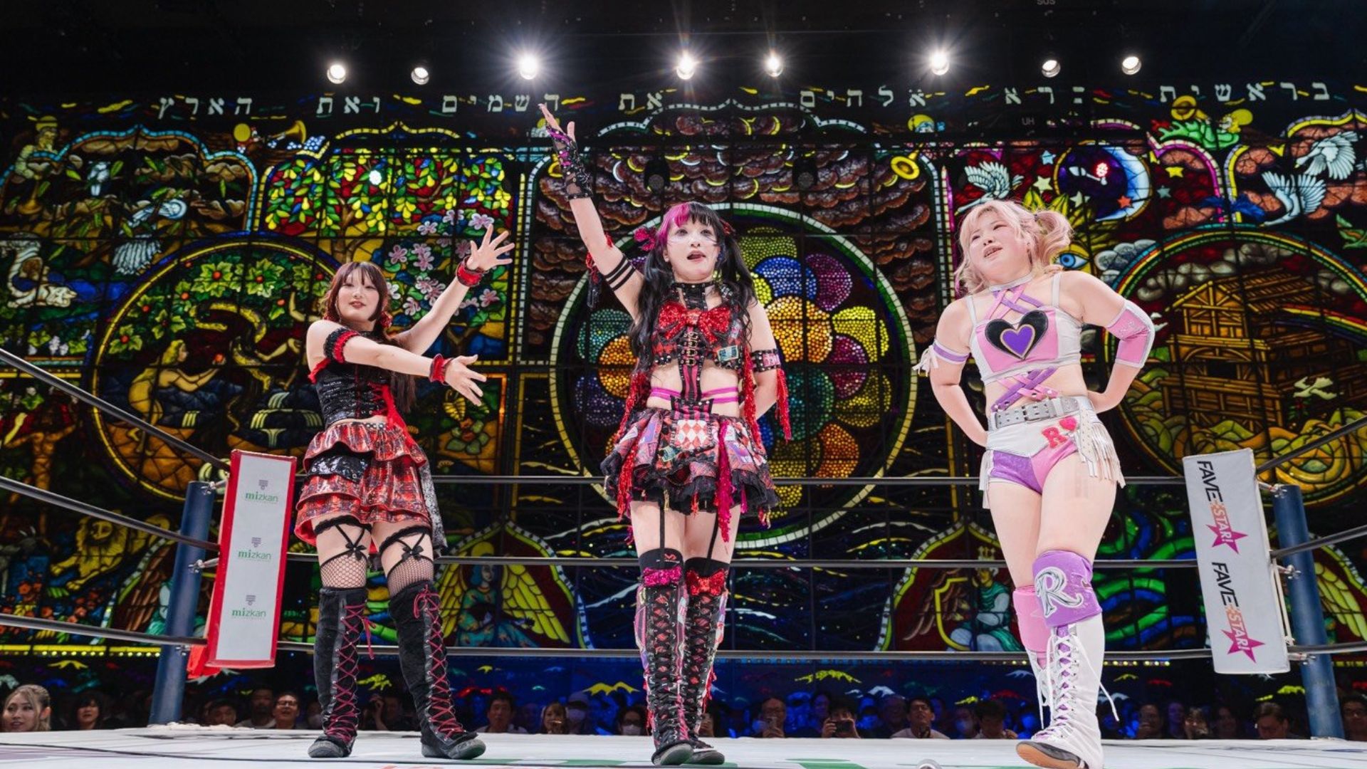 Three female wrestlers pose in a colorful ring with a stained-glass backdrop, bright costumes and dramatic gestures.