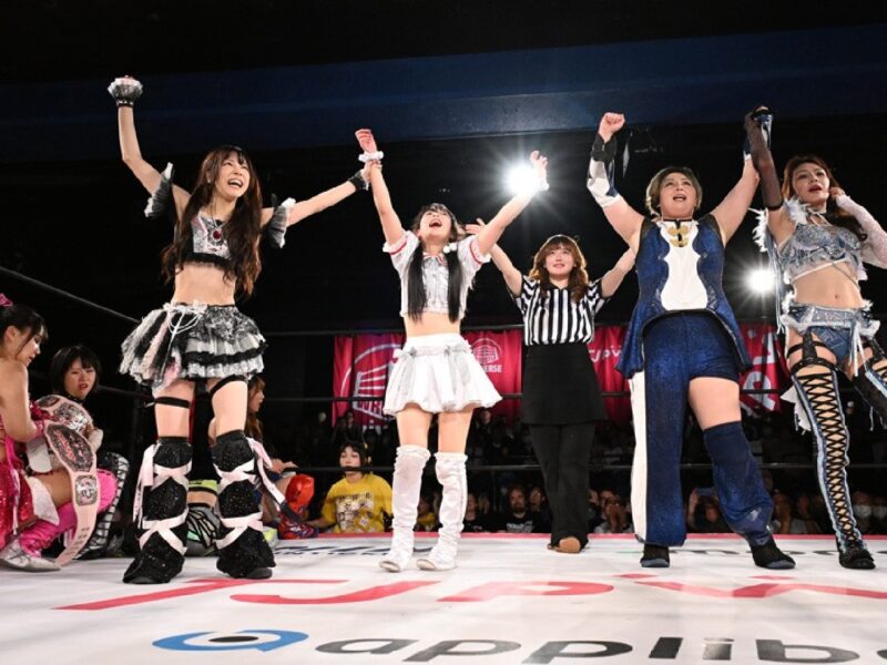Women wrestlers in colorful costumes celebrate victory in the ring, with the referee raising hands, as the crowd cheers.