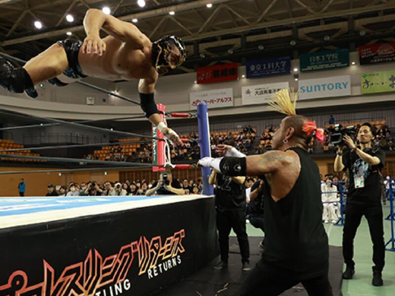 Wrestler in midair performing a high-flying dive toward an opponent inside the ring during a match, with spectators in the background on arena seating.