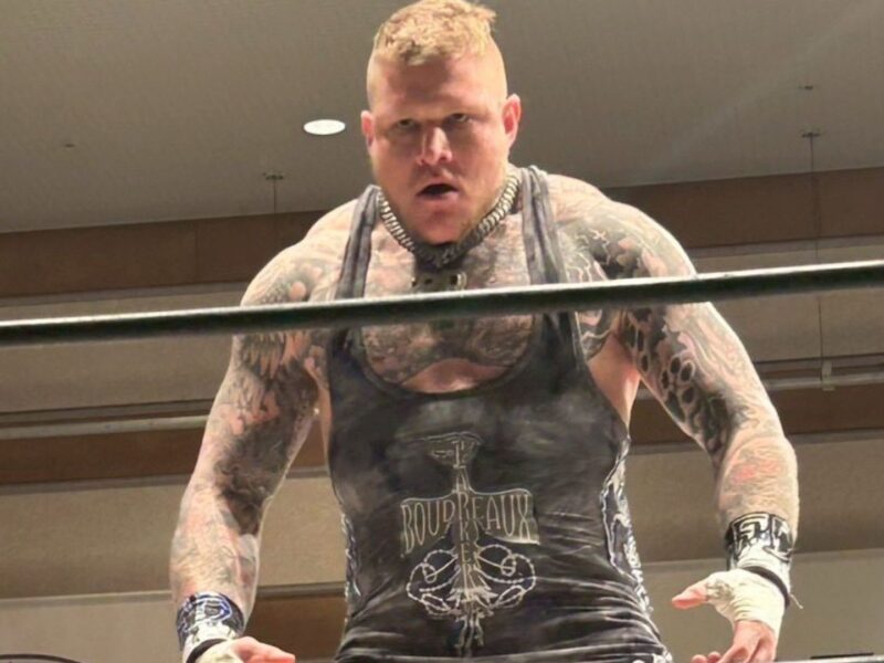 Tattooed muscular wrestler leaning on the ropes in a ring, wearing a black sleeveless singlet and a spiked collar, looking intense at the camera.