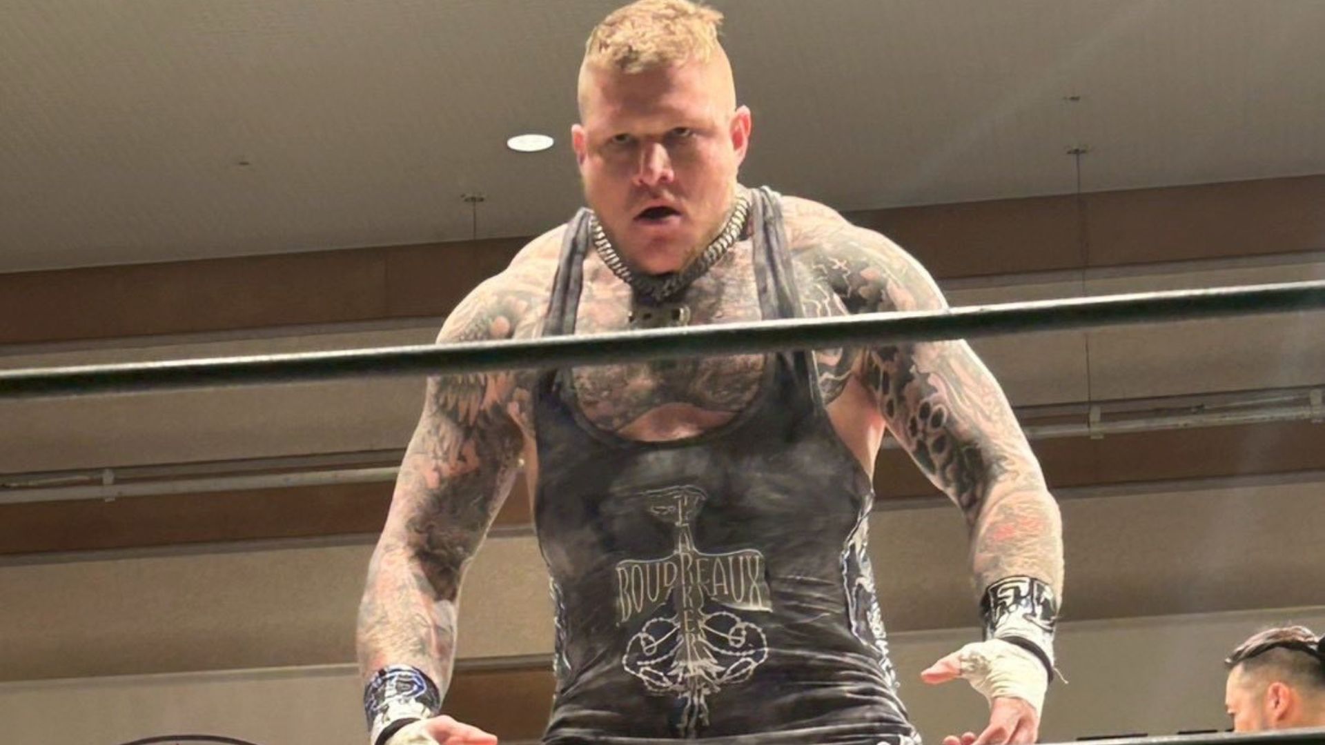 Tattooed muscular wrestler leaning on the ropes in a ring, wearing a black sleeveless singlet and a spiked collar, looking intense at the camera.