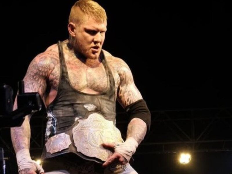 Muscular tattooed wrestler wearing a black outfit and a large championship belt, standing in a dark arena.