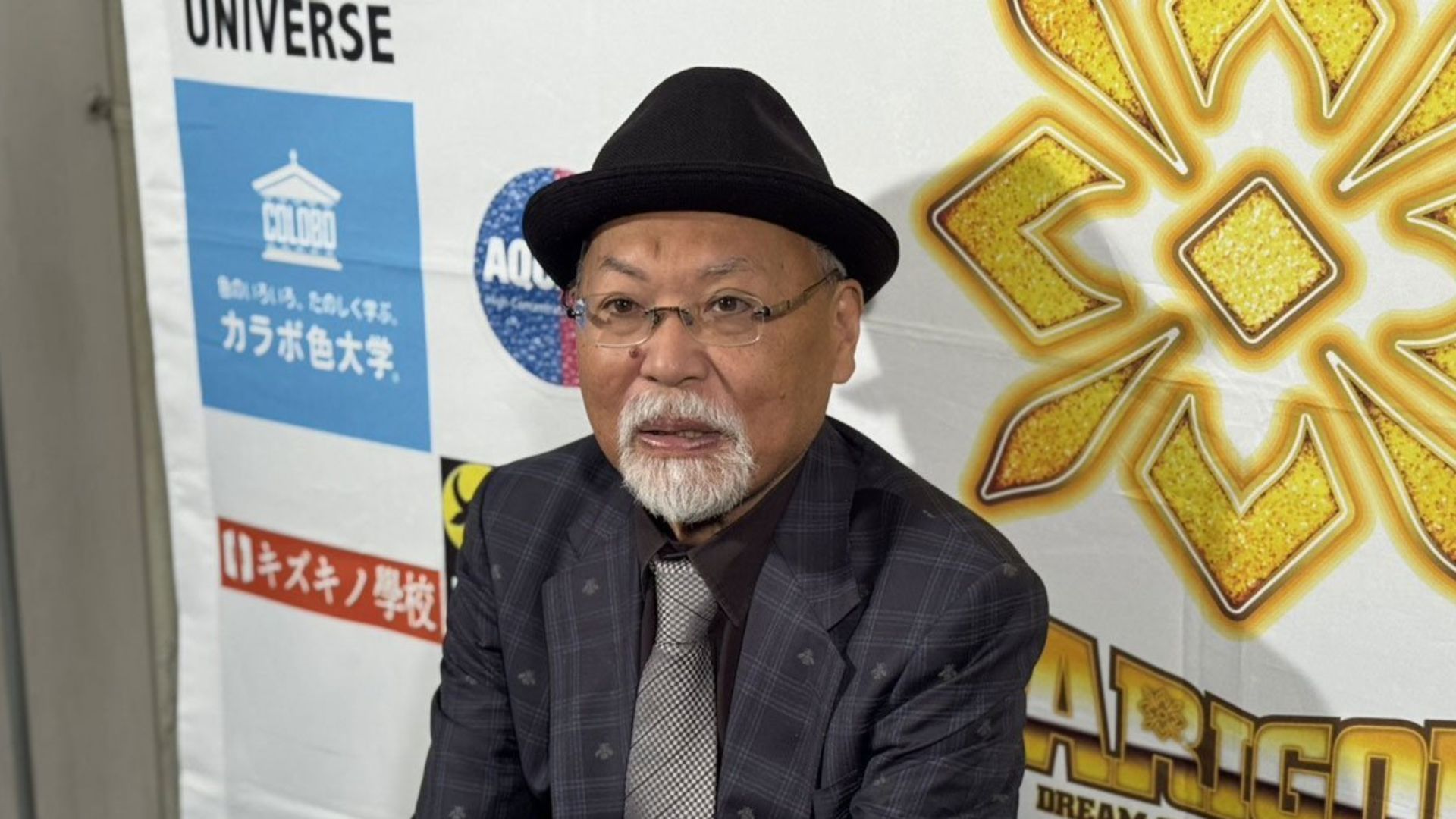 Older man with a black hat and glasses in front of a vibrant banner with Japanese writing and bold gold graphics.