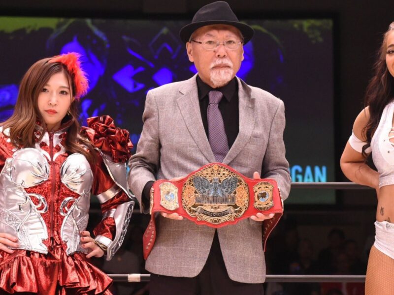 Three wrestlers pose in a ring, with the central man holding a championship belt while flanked by two women in elaborate outfits.