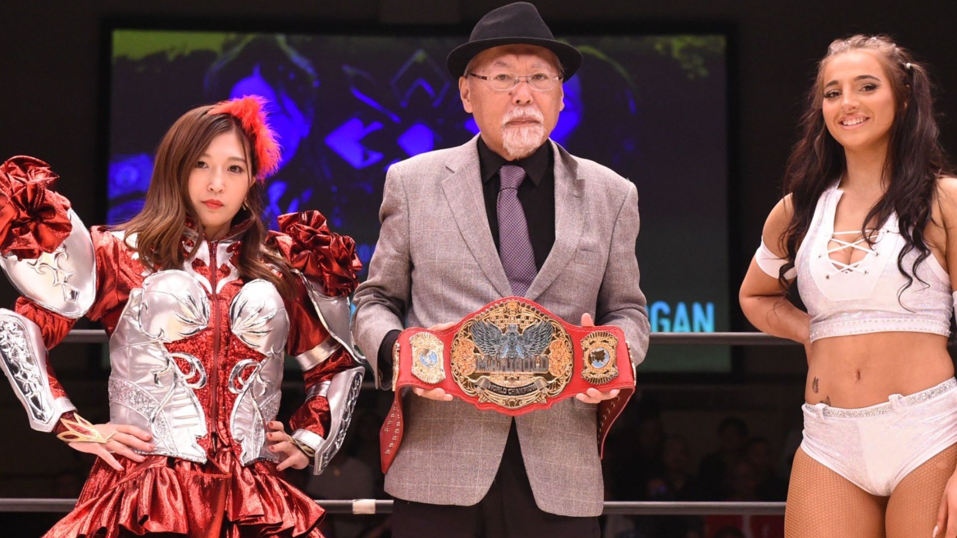 Three wrestlers pose in a ring, with the central man holding a championship belt while flanked by two women in elaborate outfits.