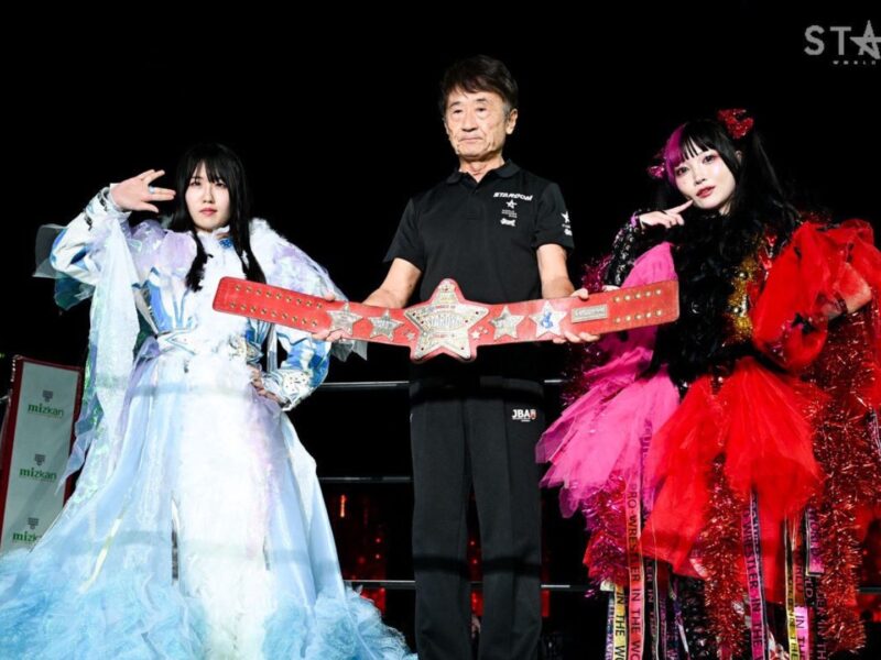 Three wrestlers pose on a black background, centerman holds a red star championship belt flanked by two in elaborate gowns.