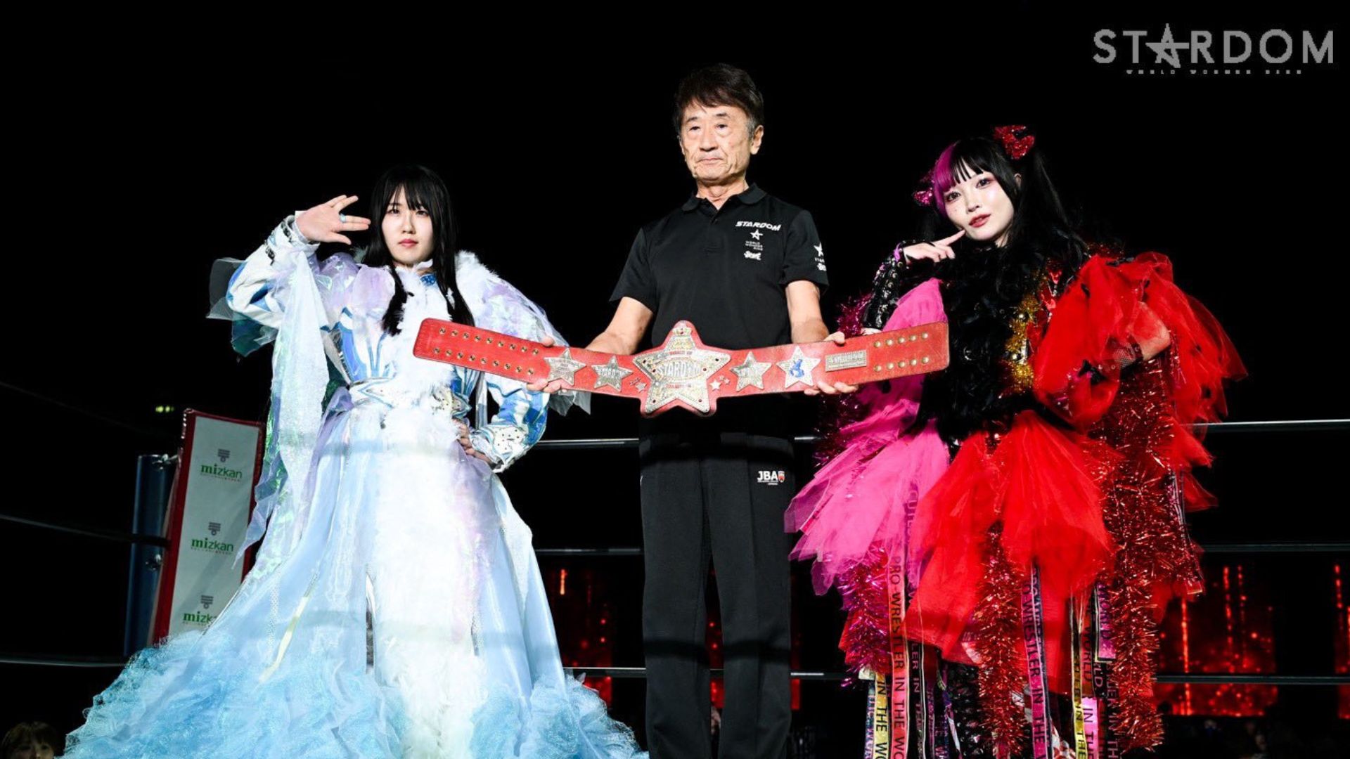 Three wrestlers pose on a black background, centerman holds a red star championship belt flanked by two in elaborate gowns.