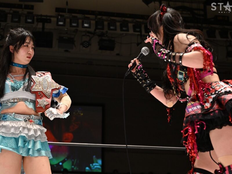 Two female wrestlers in colorful costumes on stage; one holds a jeweled championship belt while the other leans in with a microphone.