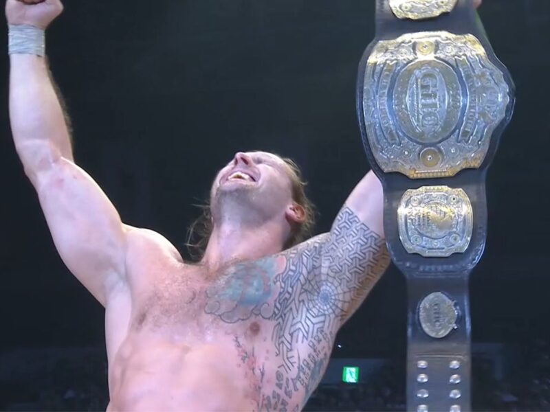 Wrestler celebrating victory, shirtless and tattooed, arms raised with a large championship belt overhead in a ring setting.