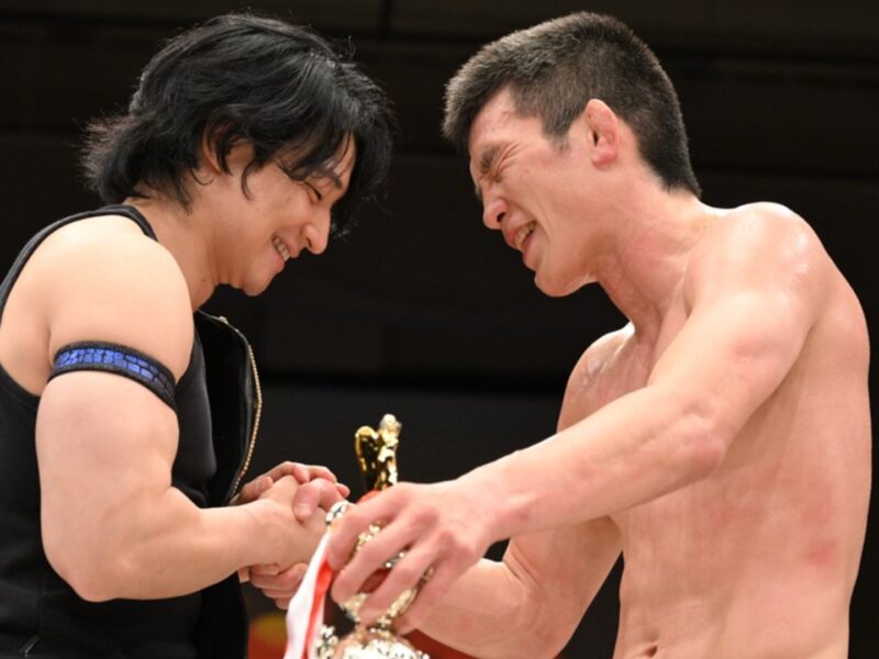 Two athletes shake hands and smile during a trophy ceremony on stage, one shirtless and the other in a black sleeveless top.