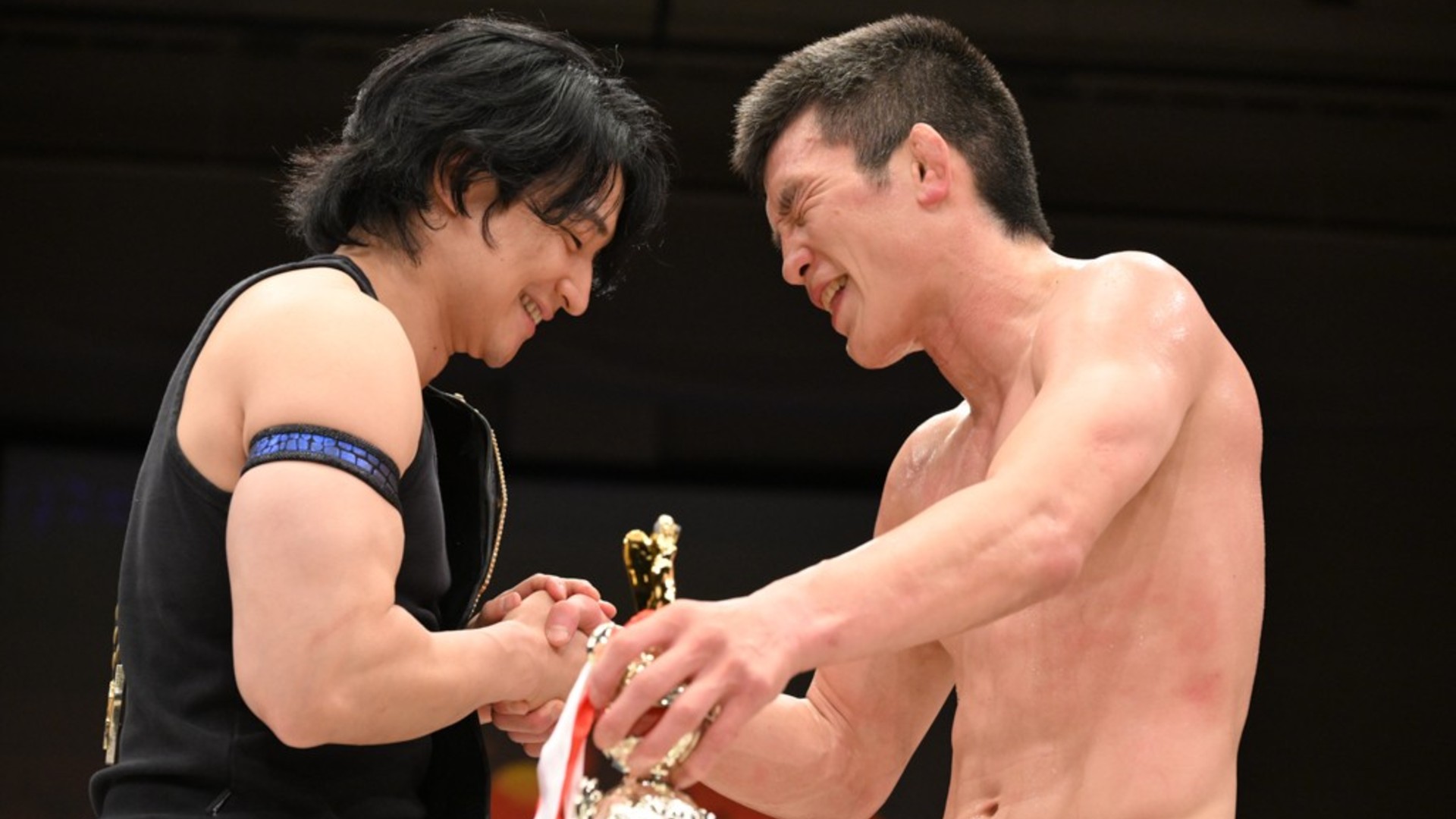 Two athletes shake hands and smile during a trophy ceremony on stage, one shirtless and the other in a black sleeveless top.