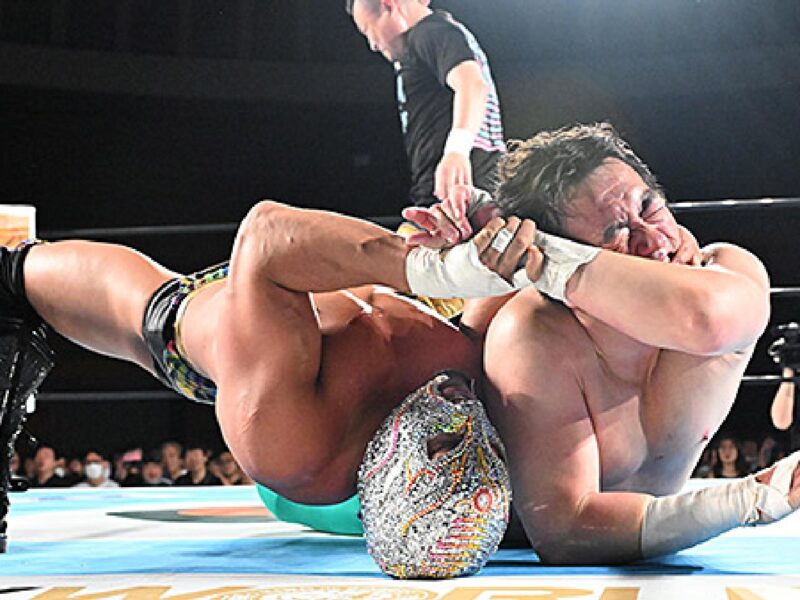 Two muscular wrestlers grapple on the mat during a match as a referee watches from the edge of the ring, crowd in the background.