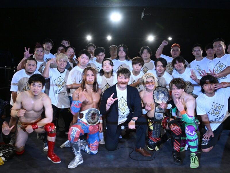 Group photo of a large pro-wrestling team in a ring, front row featuring champions with belts and two shirtless wrestlers in colorful gear, everyone posing together.