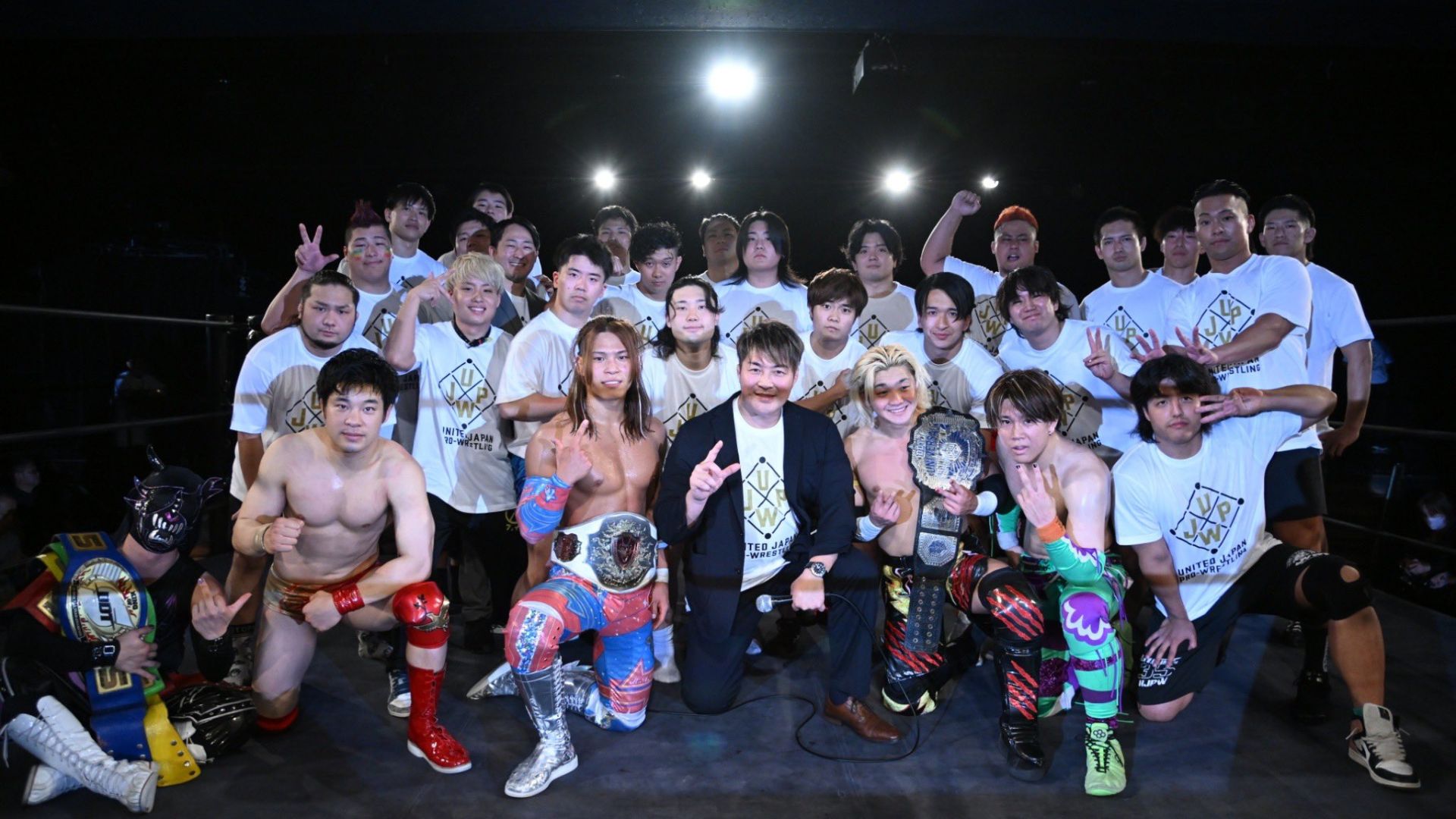 Group photo of a large pro-wrestling team in a ring, front row featuring champions with belts and two shirtless wrestlers in colorful gear, everyone posing together.