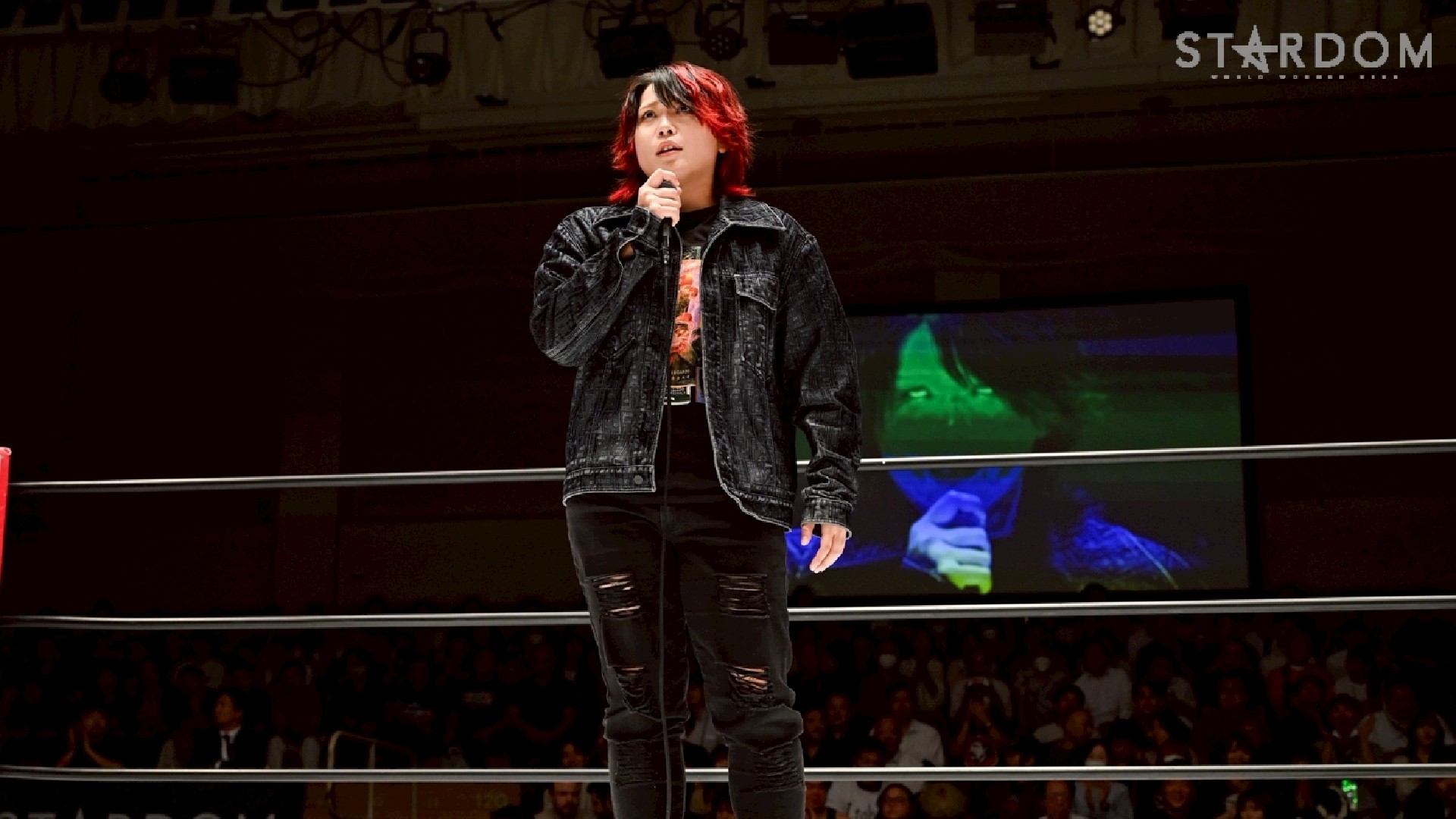 Performer with red-tinted hair speaks into a microphone on a stage inside a ring, wearing a black denim jacket and ripped jeans. A crowd sits in the dark in the background.