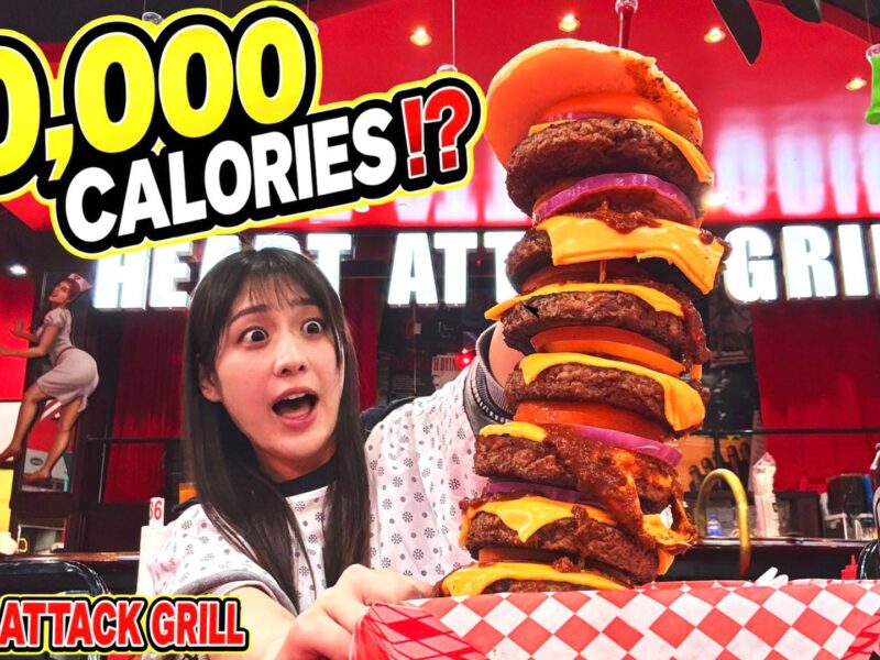 Woman holds a towering multi-patty cheeseburger at the Heart Attack Grill in Las Vegas, with bold '20,000 CALORIES' text in the background.