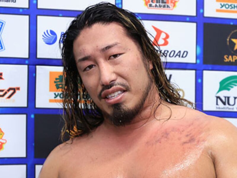 Smiling shirtless wrestler with wet hair in front of a sponsor backdrop displaying logos.